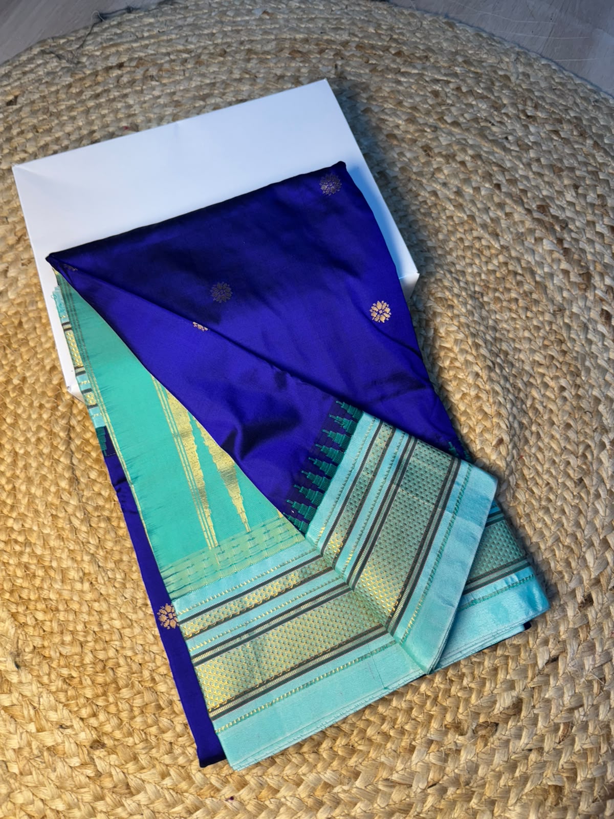 A folded silk saree displayed on a woven straw mat, featuring a deep blue-purple body with gold zari motifs and a border in mint green, teal, and gold.