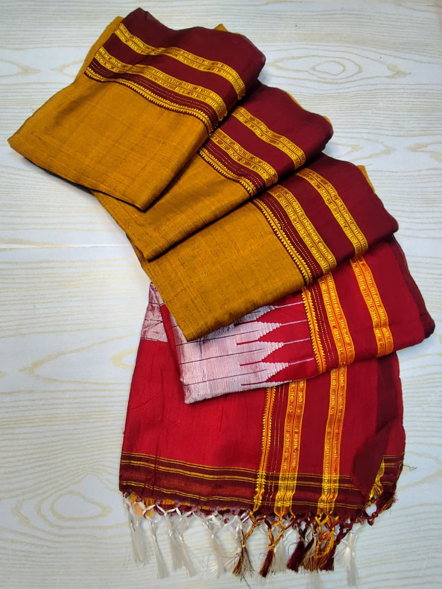 A folded traditional Indian saree or fabric set displayed on a light wood-grained surface. The fabric features a mustard yellow body, deep red borders with gold zari stripes, and a pink and red ikat patterned section with white tassels.