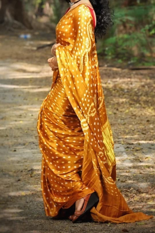 A woman wearing a golden yellow saree with white polka dots, standing outdoors on a dirt path with trees in the background.