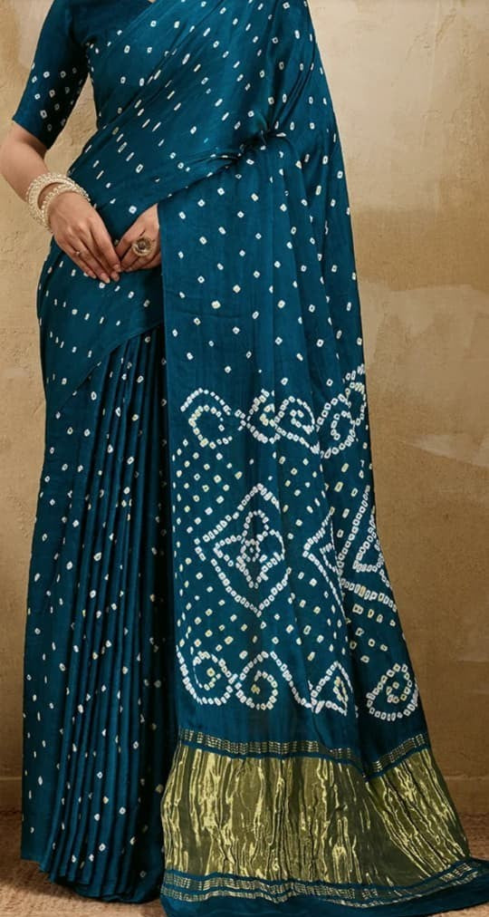 A teal blue georgette saree with white bandhani dot patterns and a metallic gold border, modeled against a neutral beige background.