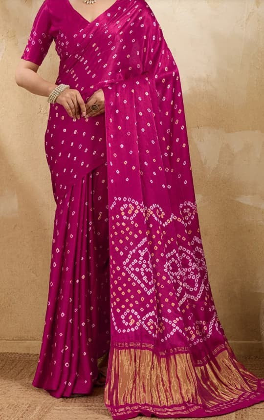 A magenta pink saree with white bandhani tie-dye patterns and a gold zari border, modeled against a textured beige background.