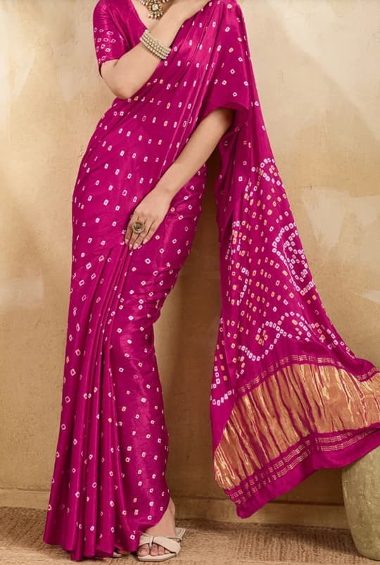 Pink Bandhani print pure modal silk saree with zari pallu detailing. A woman models a bright magenta pink saree with white tie-dye bandhani patterns and a gold zari border on the pallu, set against a textured beige background.