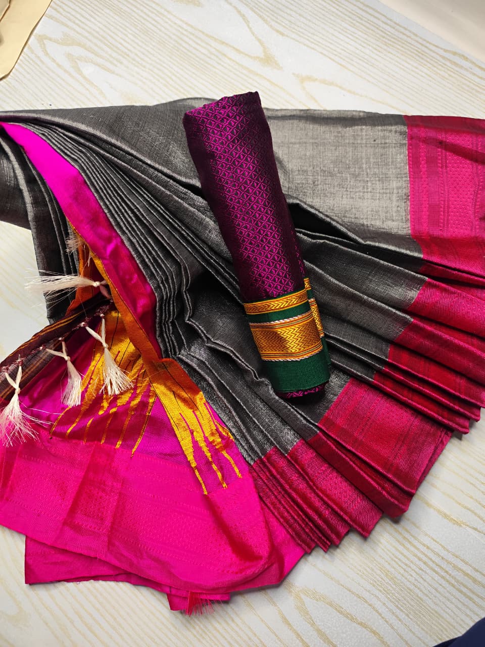 A folded and draped saree with a dark gray body, bright pink border, and a rolled purple pallu featuring gold and green zari detailing, laid flat on a light wood surface.