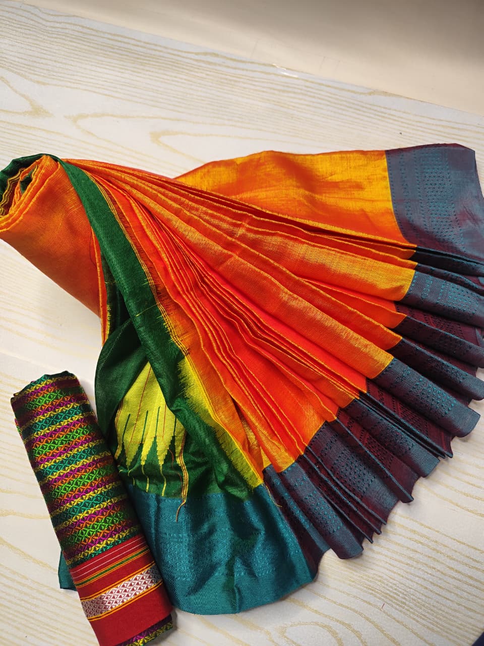 A folded and draped multicolor sari laid out on a light wood surface, with a rolled matching fabric piece placed beside it. The sari features broad bands of orange, yellow, green, teal, and dark blue with woven border details.