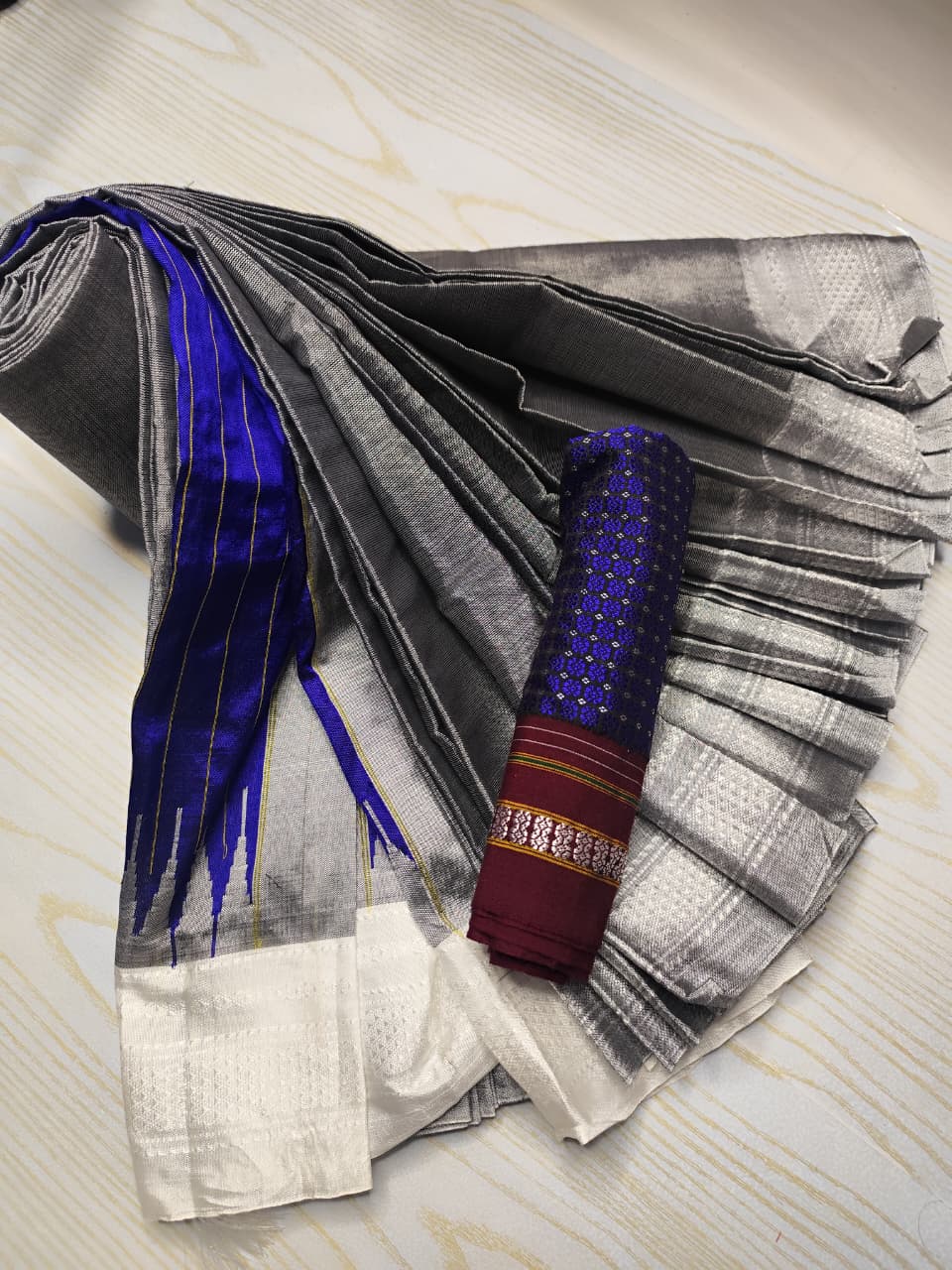 A folded and rolled traditional Indian saree displayed on a light wood surface. The saree features a silver and gray checkered body, a vibrant royal blue border with gold stripes, and a white border with a metallic silver ikat pattern.