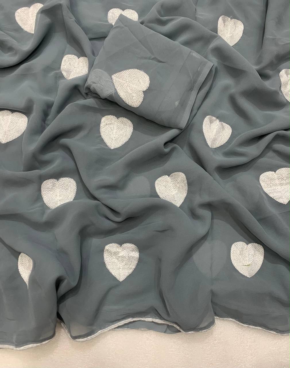 A draped piece of gray fabric with a repeating pattern of white embroidered hearts, laid out on a light neutral surface.