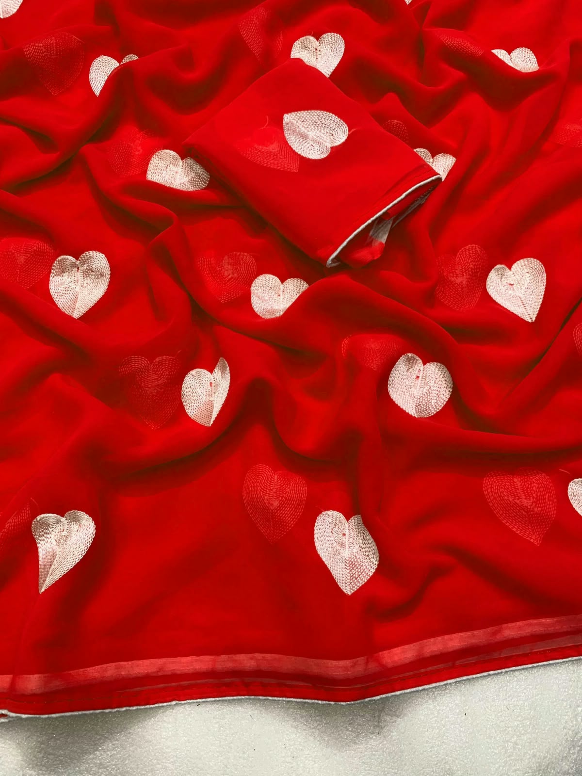 A close-up of a bright red fabric, likely a saree or dupatta, featuring scattered white embroidered hearts and a matching rolled section showing the inner border.