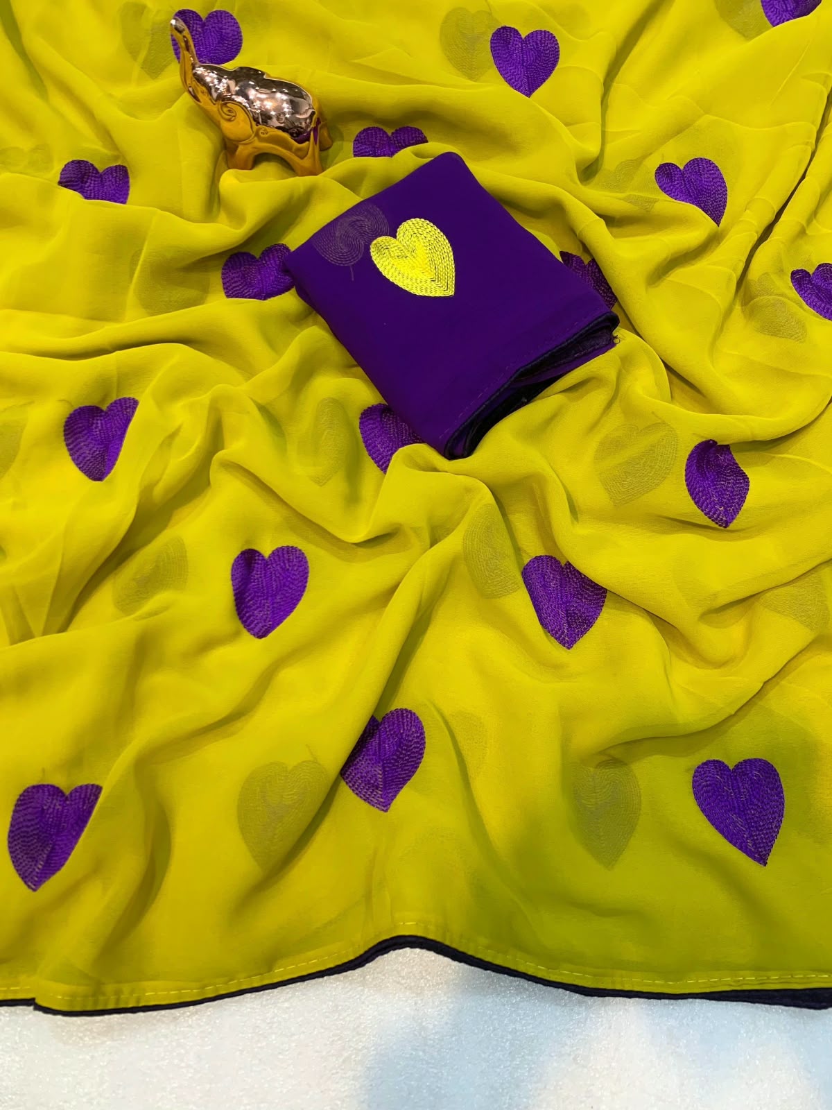 A bright yellow fabric, likely a saree, draped to show its flow and pattern of purple embroidered hearts. A matching purple fabric piece with a yellow heart is placed on top, along with a small gold elephant figurine prop.