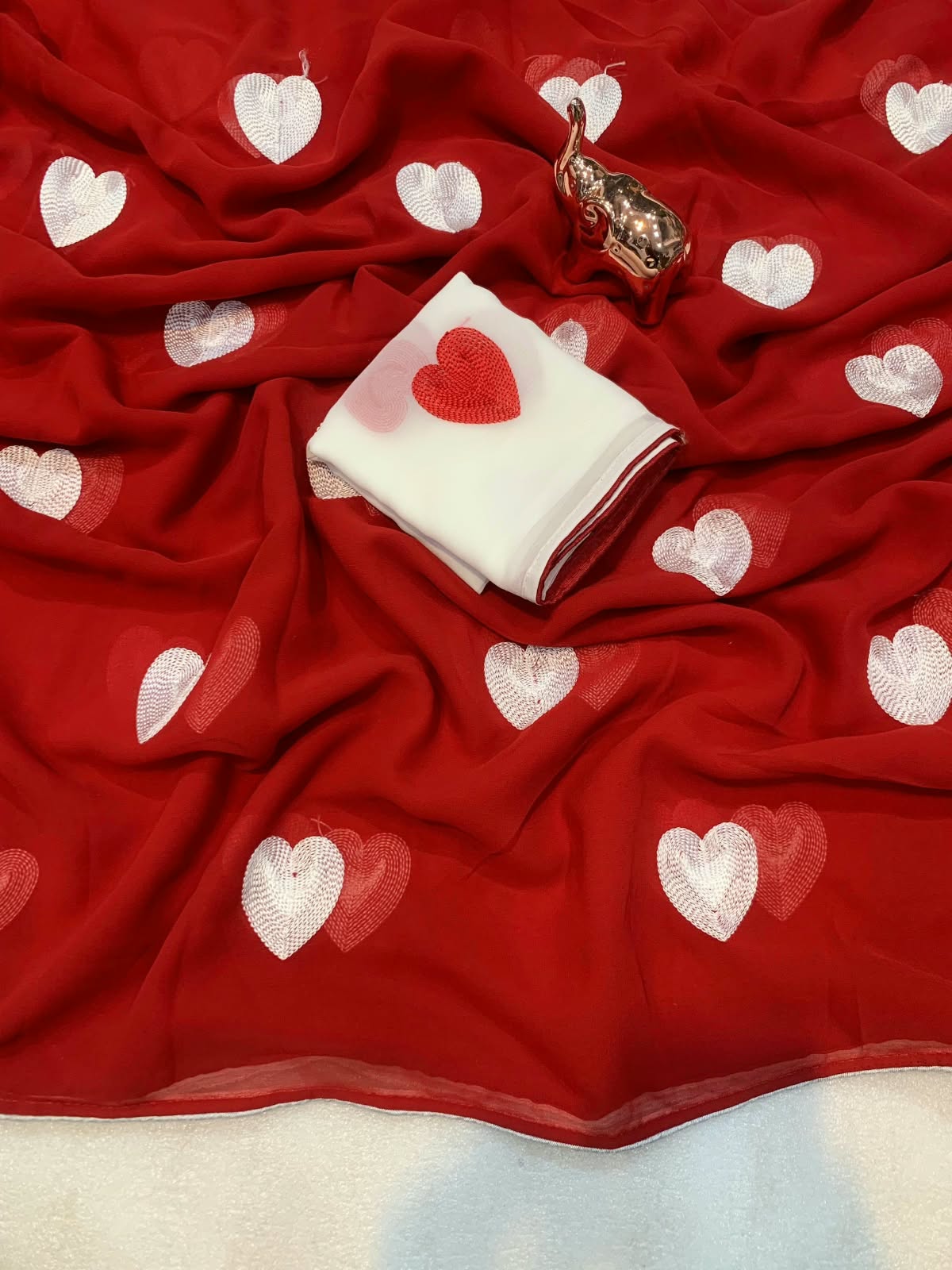 A red fabric, likely a saree or dupatta, decorated with white and light pink embroidered hearts, styled with a folded white cloth featuring a red heart and a small metallic elephant figurine.