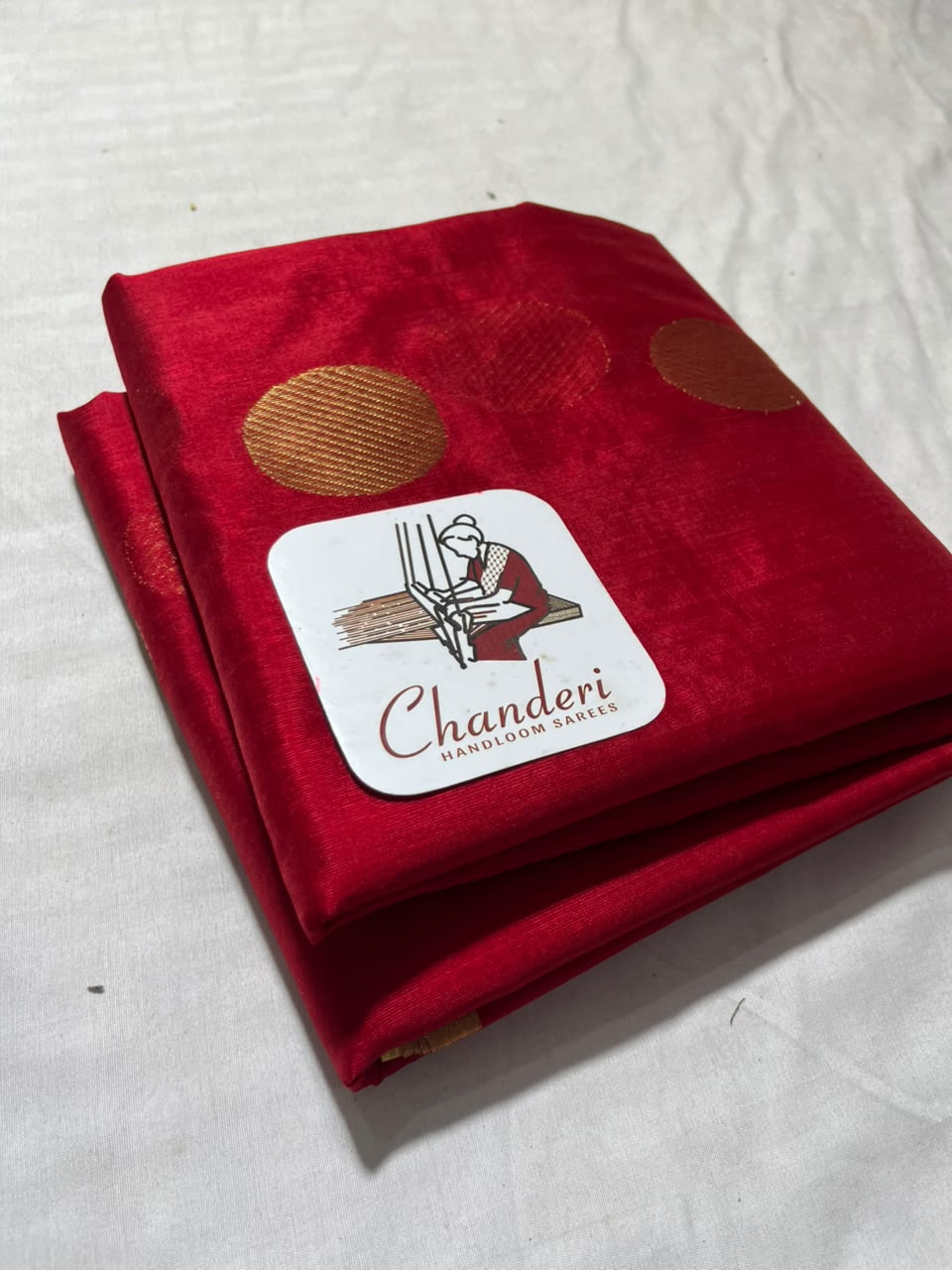 A folded red Chanderi silk fabric piece with metallic gold motifs and a white brand label reading 'Chanderi Handloom Silk' on a white textured background.