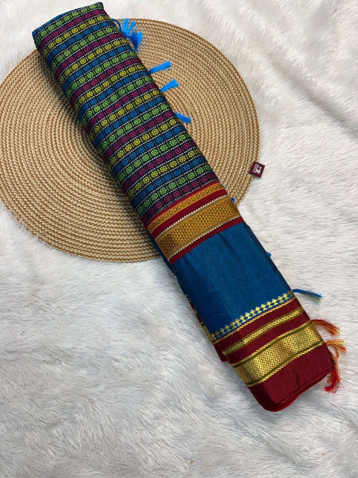 A rolled multicolor fabric item, likely a scarf or shawl, featuring blue, green, red, and gold patterns, displayed diagonally on a woven straw mat over a white faux fur background.