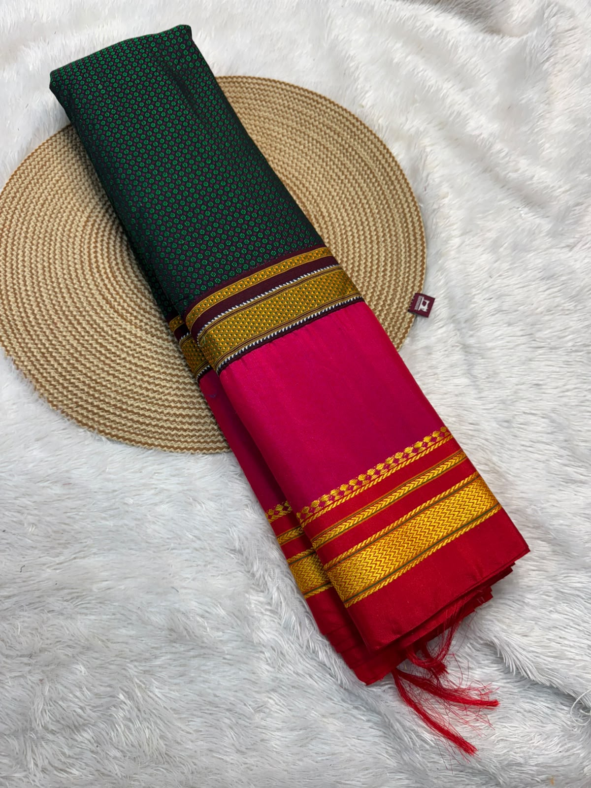 A folded traditional scarf or shawl with a dark green upper section and a bright pink lower section, decorated with gold and red striped borders, displayed on a round woven straw mat over a white faux fur background.