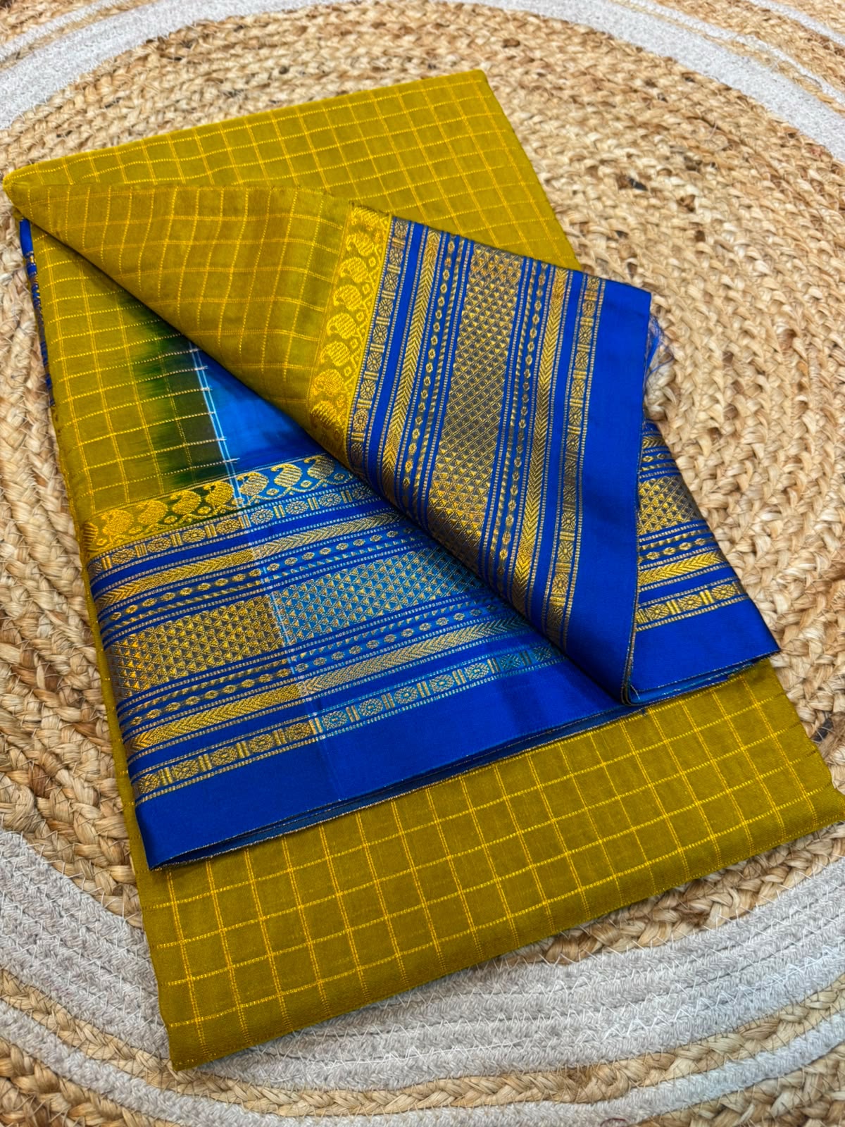 A folded yellow and blue saree with intricate gold zari patterns and borders, displayed on a woven straw mat.