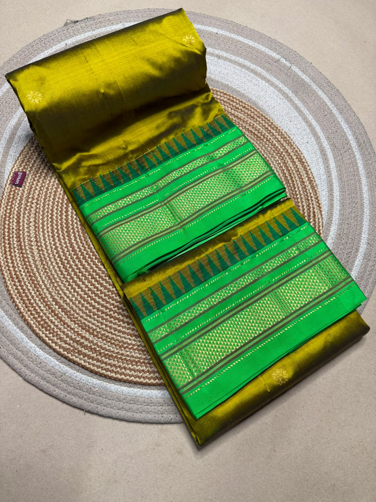 A folded and rolled green and yellow fabric saree with intricate woven patterns and borders, displayed on a circular woven placemat over a neutral surface.