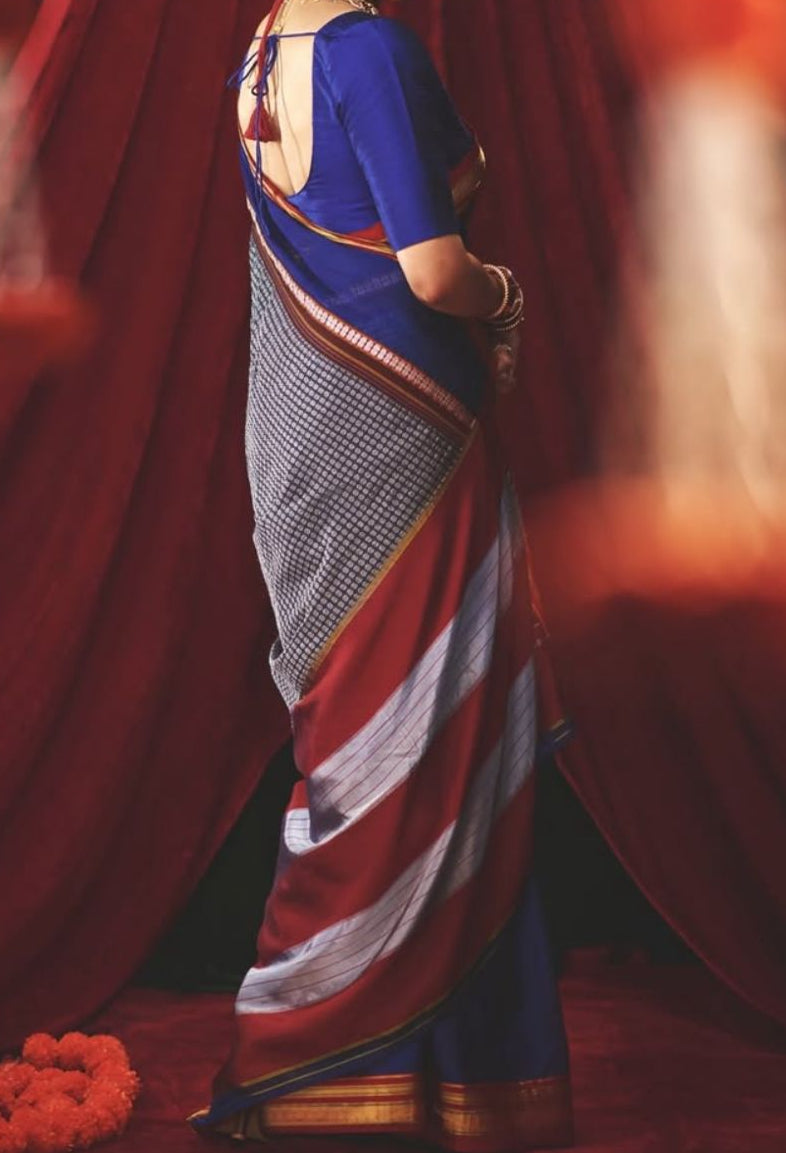 A woman wearing a traditional Indian sari stands in a studio setting with red draped fabric behind her. The sari has a royal blue upper section, a gray and white patterned body, and red and white striped lower drapes with a gold border.