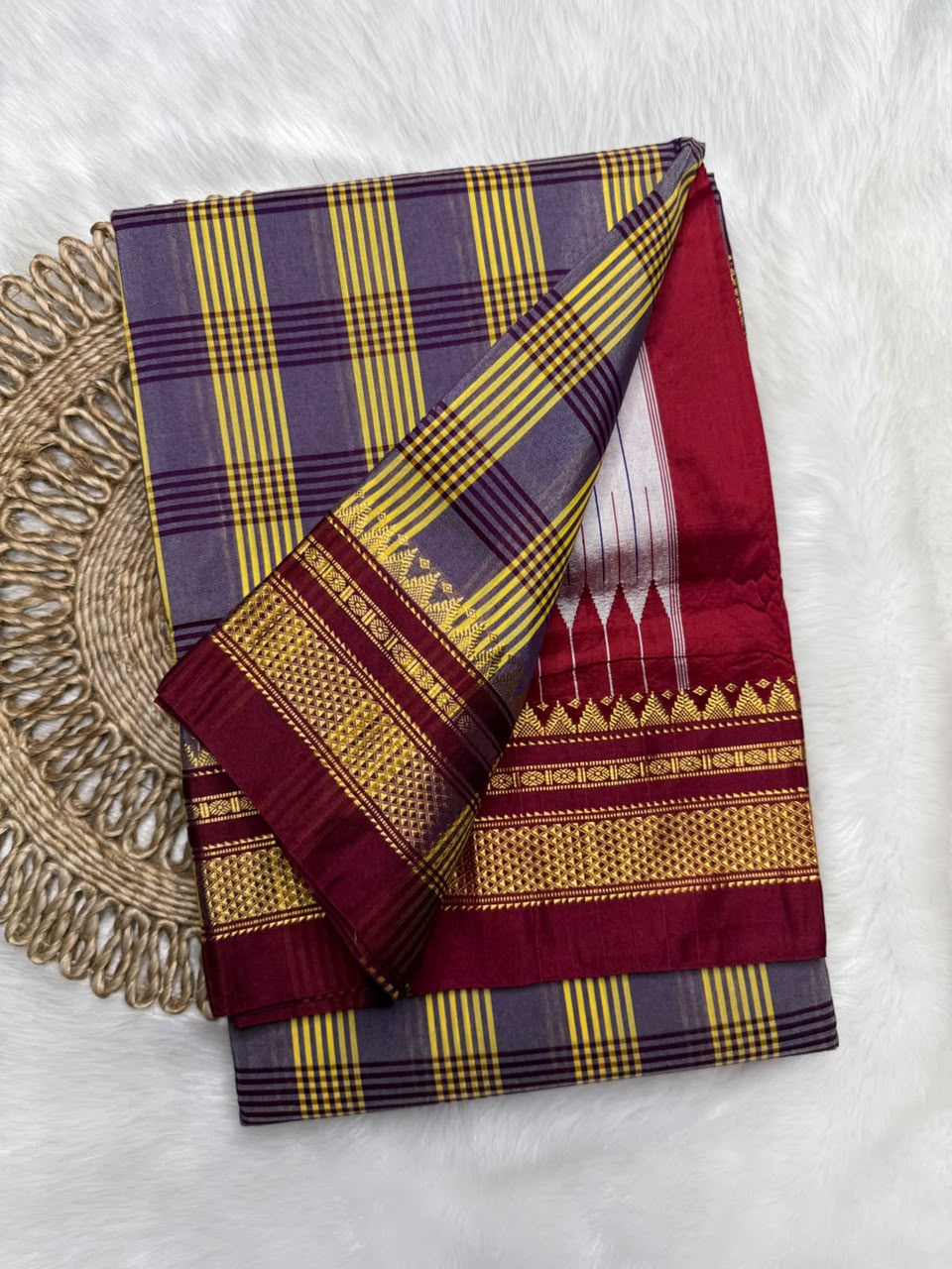A folded traditional Indian saree with a purple and yellow checkered body, a deep maroon border, and gold woven patterns, displayed on a white faux fur background next to a woven placemat.