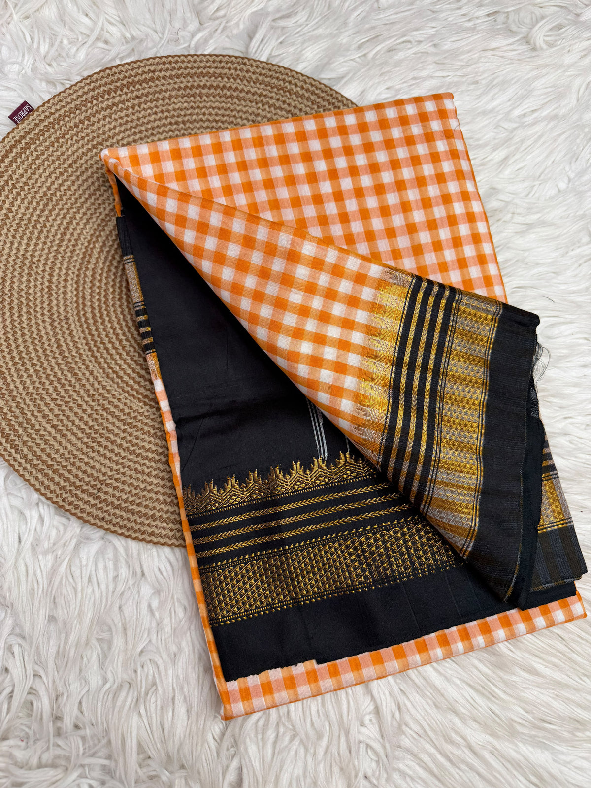 An orange and white gingham check saree with a black and gold zari border, folded on a white faux fur rug next to a round woven placemat.