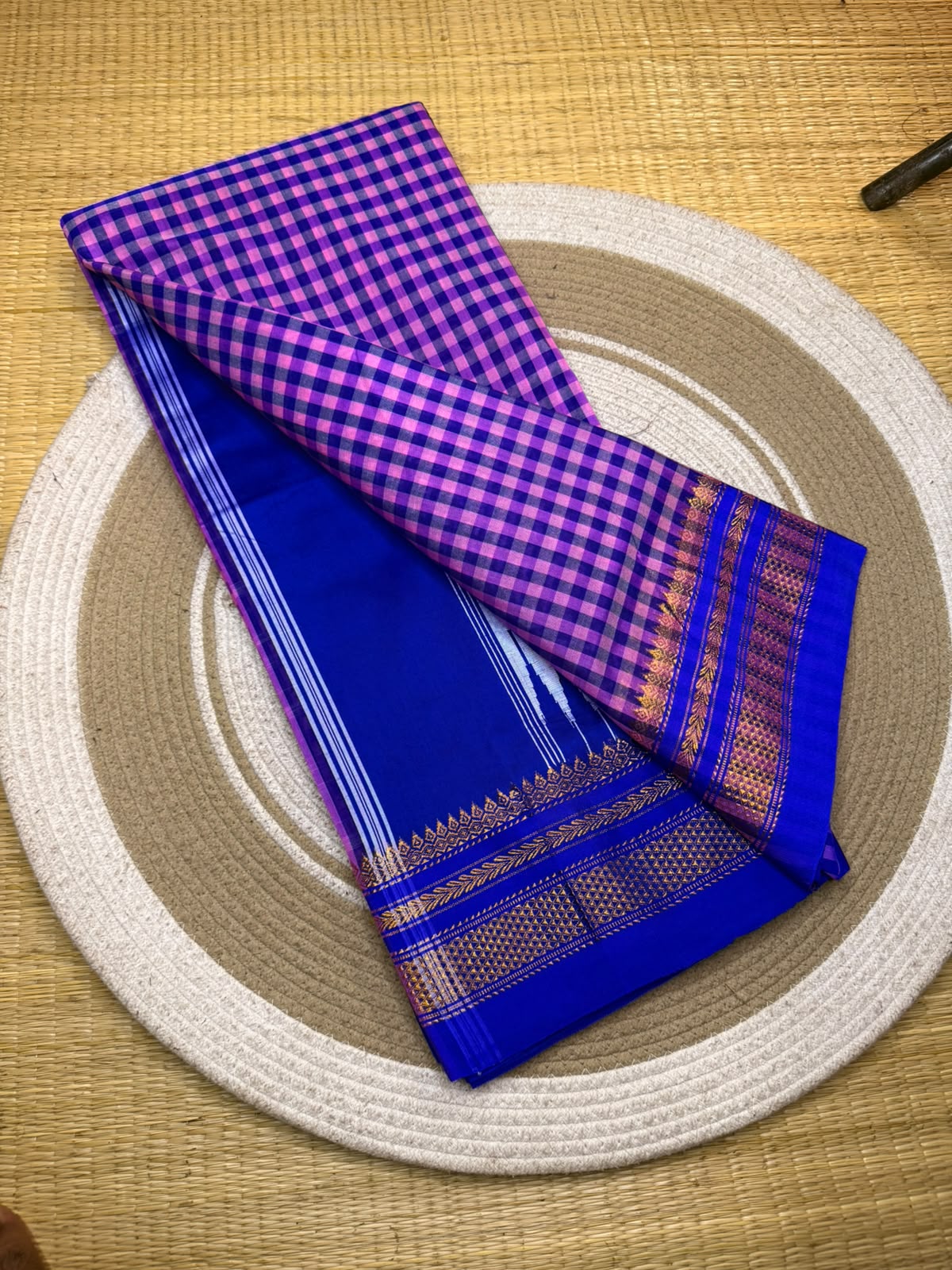 A folded royal blue and purple checkered saree with a gold zari border, displayed on a round woven placemat over a textured beige mat.