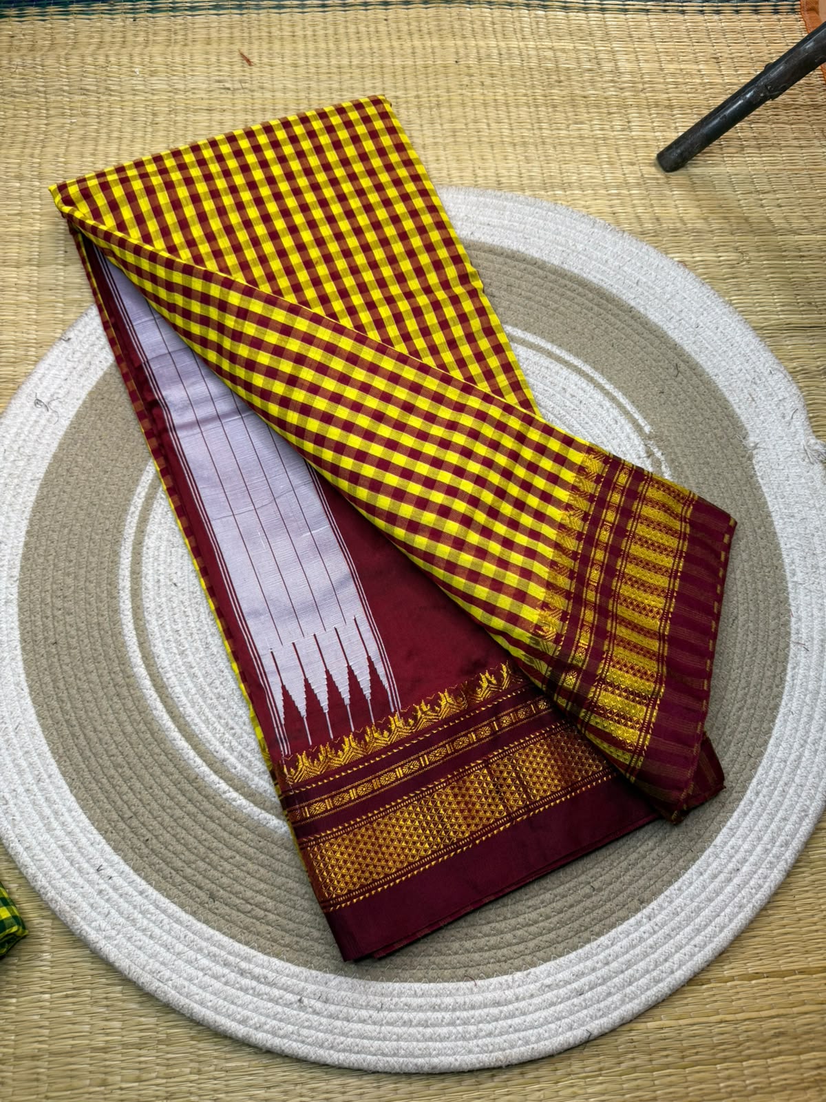 A folded saree with a yellow and maroon checkered body, a deep maroon border, and a white and maroon zari-painted pallu, displayed on a round woven mat.