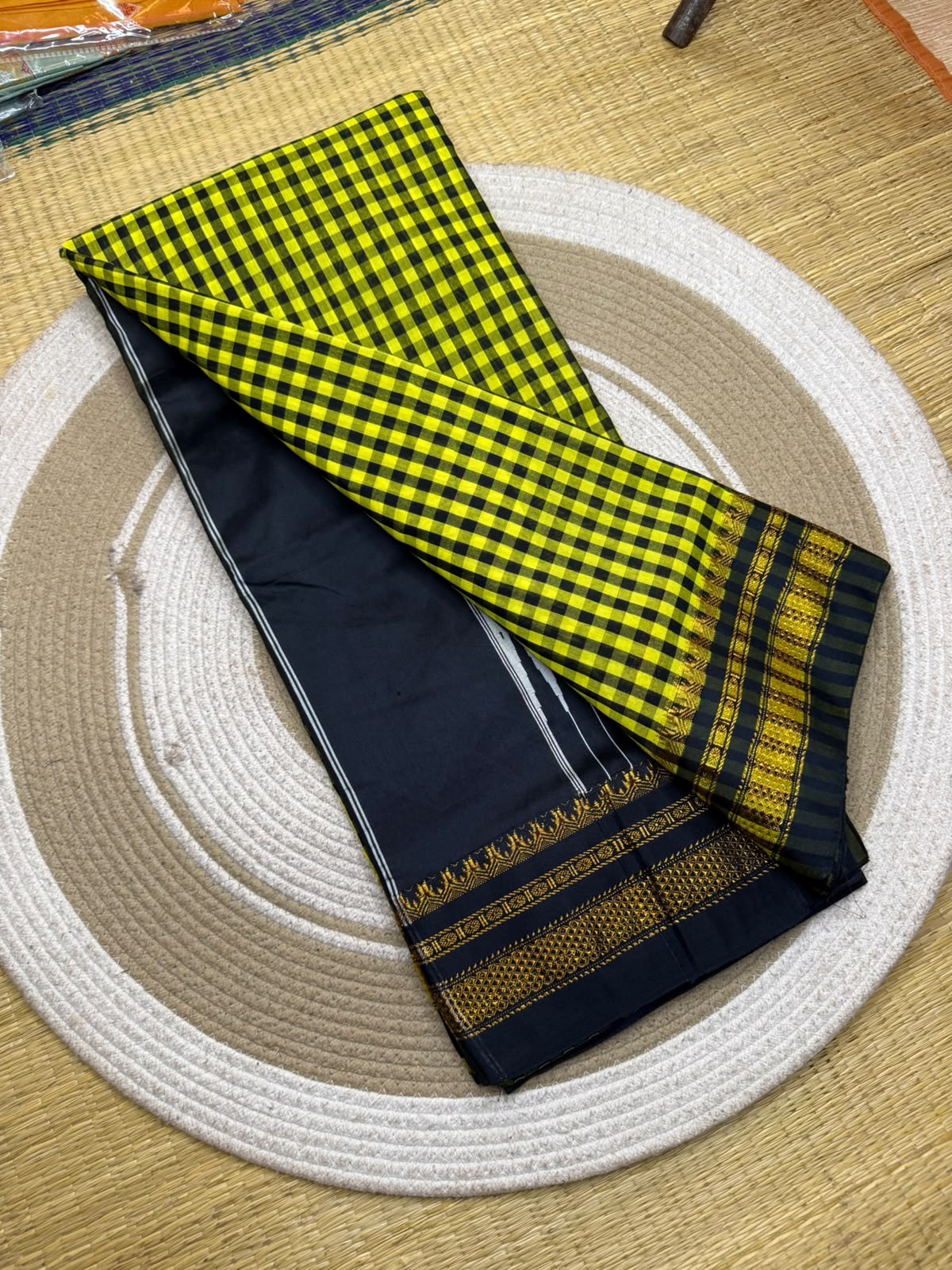 A folded black and yellow checkered fabric, likely a saree or textile piece, displayed on a round woven straw mat over a light beige rug.