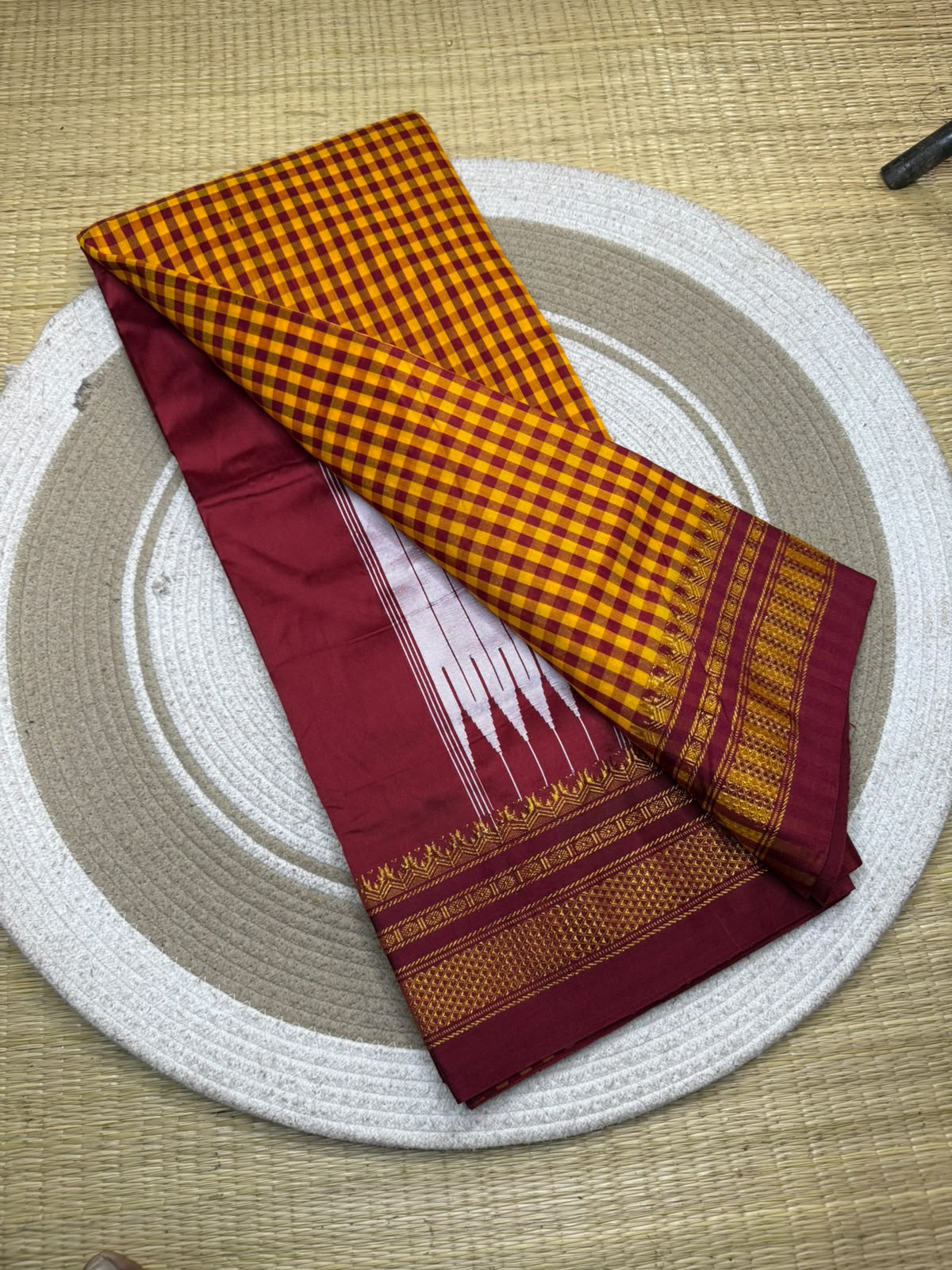 A folded traditional Indian saree displayed on a round woven mat. The fabric features a golden-yellow and red gingham check pattern on one section, a deep maroon border with white and gold motifs, and a gold-embroidered pallu.