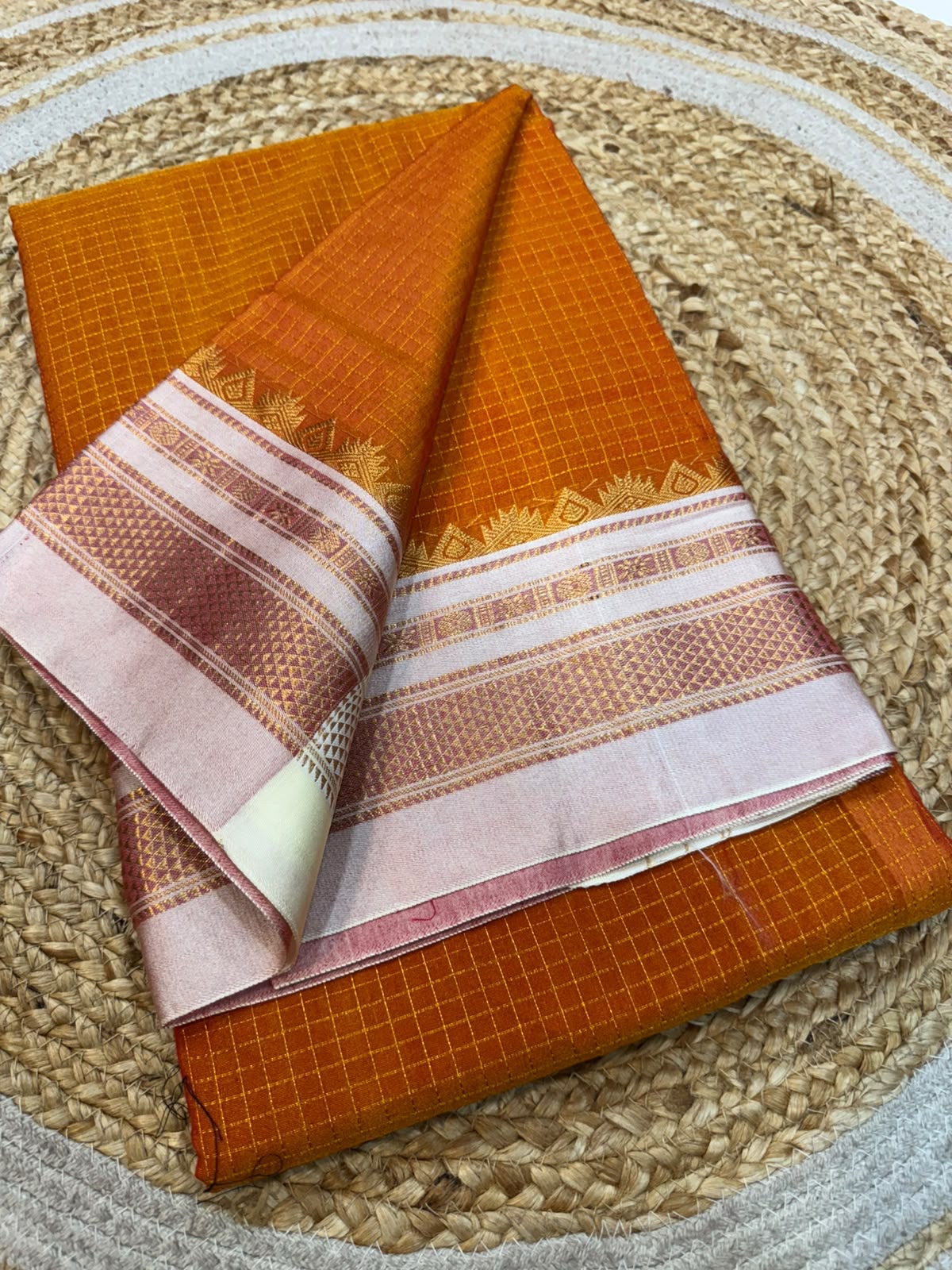 An orange woven fabric saree or textile with a pink and white patterned border, folded and displayed on a round woven straw mat.