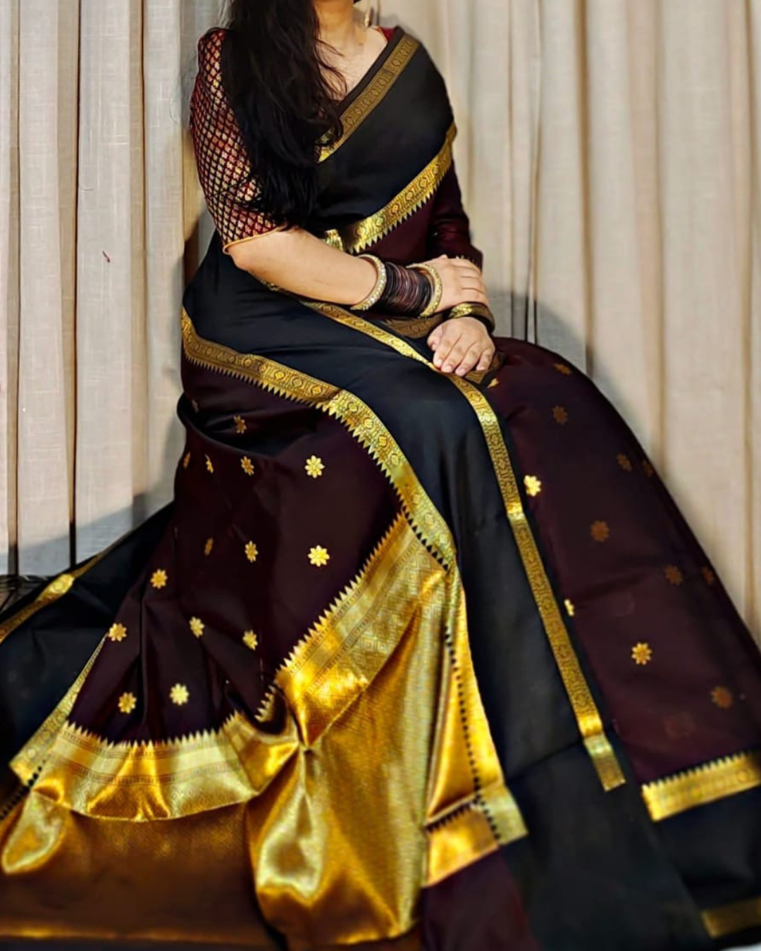 A woman seated indoors wearing a dark sari with a wide gold border, gold floral motifs, and a matching gold blouse. The background is a beige curtain.