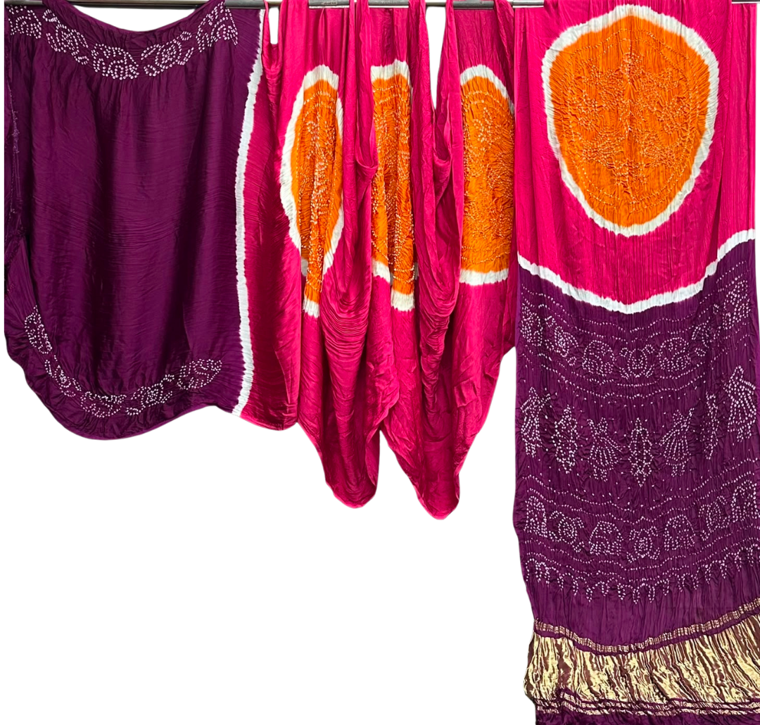 Three colorful traditional draped garments or fabric pieces hanging against a plain white background. The fabrics feature bold patterns including orange circular tie-dye sections, white border details, and purple embroidered borders.