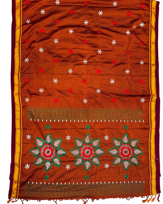 A rust-orange woven textile, likely a traditional scarf or sari, featuring embroidered floral motifs, star patterns, and geometric borders in red, green, white, and yellow.