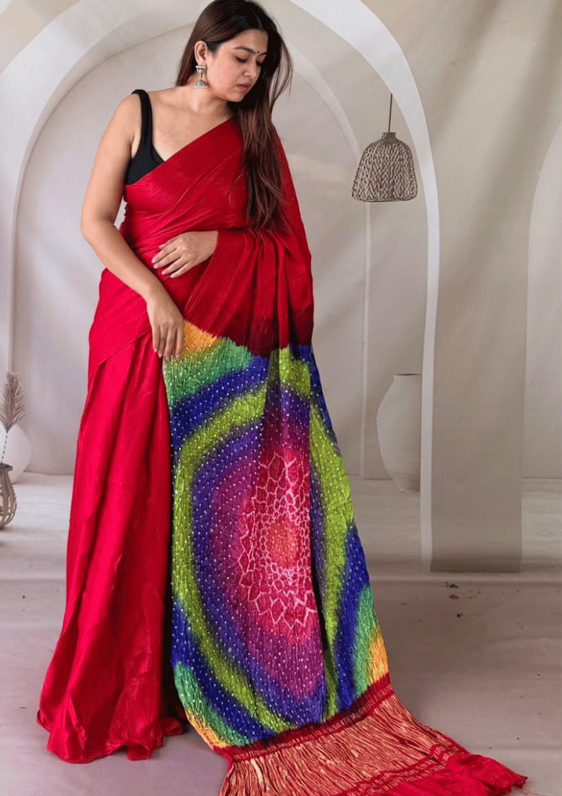 A woman models a vibrant red saree with a multicolored rainbow spiral pattern on the pallu and a red fringed border, paired with a black sleeveless blouse, set against a neutral arched interior background.