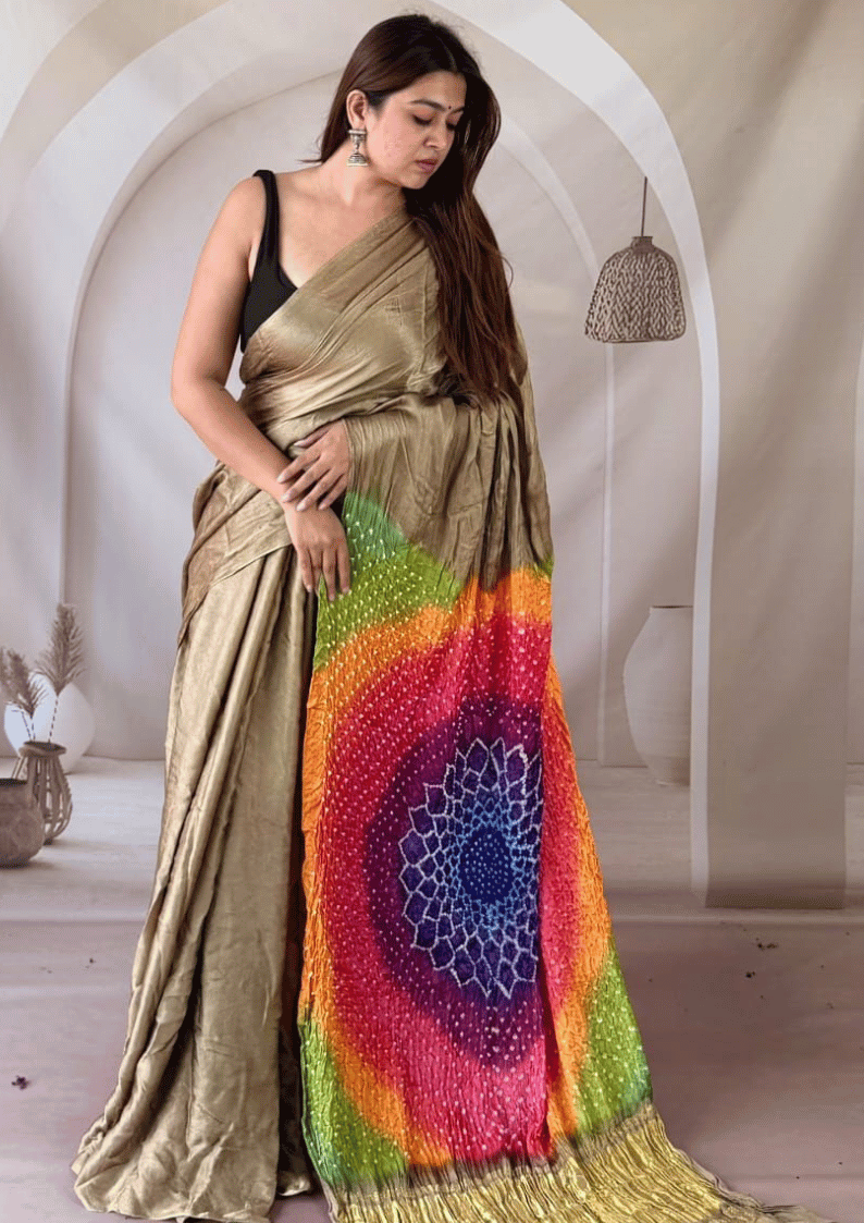 A woman models a metallic gold saree with a vibrant multicolor mandala print on the lower portion, paired with a black sleeveless blouse, set against a neutral arched interior background.